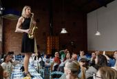 Eine der Musikerinnen auf einem Tisch stehend, während sie Saxofon spielt, umgeben von begeisterten Zuschauern