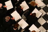 Vista aérea de varios miembros de un coro durante una actuación. Los cantantes, vestidos con ropa formal negra, están de pie sosteniendo carpetas de música abiertas. Se encuentran en la Real Basílica de Nuestra Señora de Atocha en Madrid, con suelo de baldosas en blanco y negro, mientras revisan sus partituras.