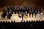 El coro en un gran escenario, vestido de oscuro, con un público frente a ellos